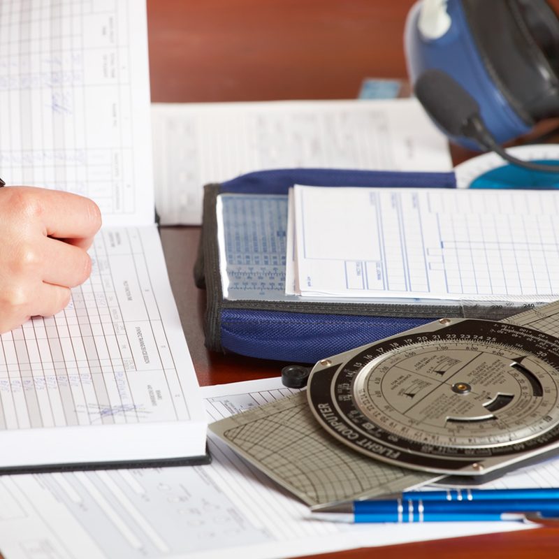 Pilot equipment with airplane pilot hand filling in logbook, other tools like flight computer used for aviation calculations, protractor, kneepad with charts and professional headset